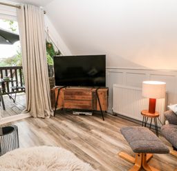 A living room with a television on a wooden stand a lamp on a side table and a chair with ottoman at Lavender Lodge in St Ann's Chapel Cornwall