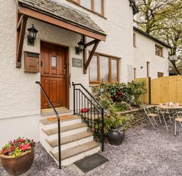 The entrance of a white cottage with wooden door and steps leading up to it with outdoor table and chairs nearby at Rose Cottage Burneside