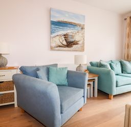 A room with two blue sofas a side table with baskets and lamps and a painting of a boat on the wall at 16 Ocean Heights in Newquay