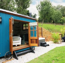 A blue corrugated metal tiny house with open wooden doors showing a white armchair inside a green grassy yard with trees and a golf cart at Little Silver Pheasant in High Bickington