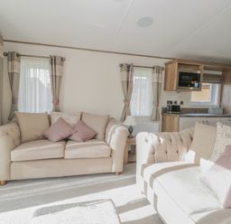 A living room with two beige sofas with cushions a small wooden side table with a lamp and a kitchen area with cupboards a microwave and a toaster at 22 Sherwood South Lakeland Leisure Village near Warton