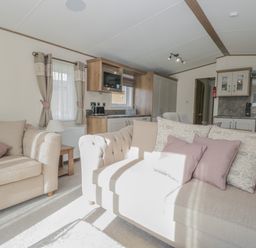 A living room with beige sofas and pink cushions next to a kitchen area at 22 Sherwood South Lakeland Leisure Village near Warton