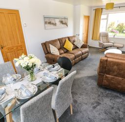 A living room with brown sofas a glass dining table set for six and a large window with yellow curtains at 53 St Brides View in Roch