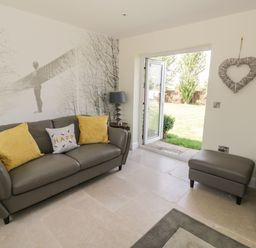 A living room with a gray sofa with yellow pillows a small table with a lamp an ottoman and an open door to a garden at Garden Cottage in Upton Upon Severn