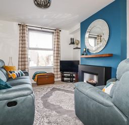 A living room with teal sofas cushions a TV on a corner stand a fireplace with a round mirror above and checked curtains at Penmar in Looe