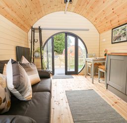 An interior view of a wooden pod with a black sofa with pillows on the left side a dining table with chairs on the right and a circular glass door at the far end at Filbert Pod in Arundel West Sussex