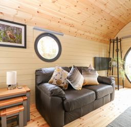A living room with a black leather sofa with cushions nest of tables a lamp plant a TV and round windows at Filbert Pod in Arundel West Sussex
