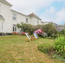 A garden with two wooden deck chairs and a small table in front of a white house with flowers and shrubs at Chynance in Porthcurno