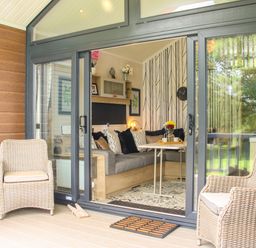 A seating area with wicker chairs outside sliding glass doors leading to a living room with a corner sofa and a table at Jacob's Meadow Pod 2 in Minsterley near Chirbury