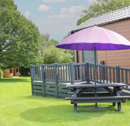 A picnic table with a purple umbrella next to a wooden building with a ramp and a grassy yard with trees and a small wooden shelter in the background at Jacob's Meadow Pod 2 in Minsterley near Chirbury