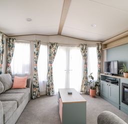 A living room with a grey sofa pink cushions patterned curtains a coffee table a TV and plants at Burlees Nest in Brigham near Great Broughton Cockermouth
