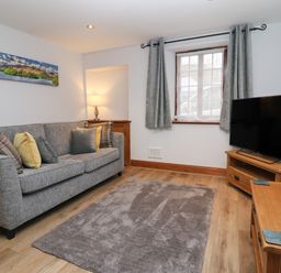 A living room with a gray sofa and cushions a wooden TV stand with a television a wooden corner cabinet a window with gray curtains and a floor lamp at Fryston Cottage in Keswick