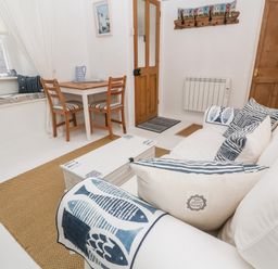 A sitting area with a white sofa and striped cushions a wooden table with two chairs and a window seat at The Little Net Loft in Mousehole near Penzance