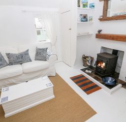 A living room with a white sofa with patterned cushions a white coffee table a wood stove fireplace with wood and tools and a wooden chair at The Little Net Loft in Mousehole near Penzance