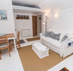 A living room with a white sofa with patterned cushions a wooden table with two chairs a white chest used as a coffee table and a floor lamp at The Little Net Loft in Mousehole near Penzance