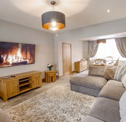 A living room with a large grey sofa a wooden TV stand a wall mounted TV showing a fireplace and a dining area with a wooden table and chairs at Family House in Weymouth