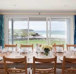 A dining room with a wooden table set with white plates glasses and a flower vase by a large window with blue curtains overlooking the sea at The White House in Polzeath
