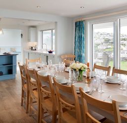 A dining area with a wooden table set with white dishes and glasses next to a kitchen with blue island and large windows at The White House in Polzeath