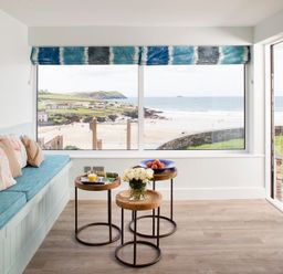 A seating area with a blue cushioned bench and three wooden round tables near large windows overlooking a beach at The White House in Polzeath