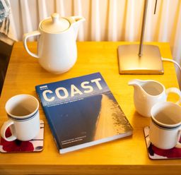 A wooden side table with a teapot two mugs a milk jug a lamp and a book titled Coast at Pencreek Rock