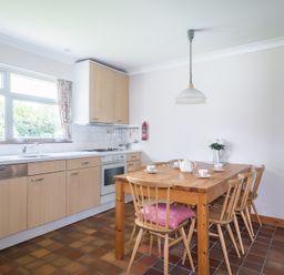 A kitchen with wooden cabinets a dining table with chairs and a window with floral curtains at Rosevallen in Daymer Bay