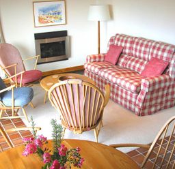 A living room with a red and white checkered sofa, wooden chairs with colored cushions, a round wooden table with flowers, a floor lamp, and a wall-mounted fireplace at Rosevallen in Daymer Bay
