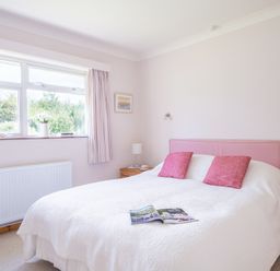 A bedroom with a bed with pink pillows and a white cover near a window with curtains and wooden bedside tables at Rosevallen in Daymer Bay
