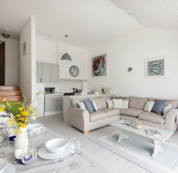 A dining table set with white dishes and a vase of yellow flowers next to a seating area with a beige sectional sofa and a coffee table at Slipway 10 in Rock