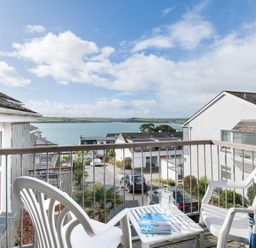 A balcony with two white plastic chairs and a small table with books and a glass bottle overlooking a street and water at Slipway 19 in Rock
