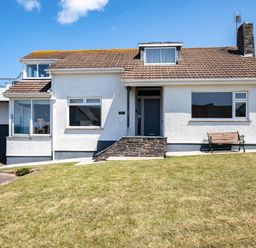 A white house with a tiled roof a front door a bench and a grassy lawn under a blue sky at Little Belz in Polzeath