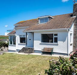 A white house with a tiled roof a bench on the front patio and a garden with flowers at Little Belz in Polzeath