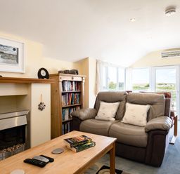 A living room with a leather sofa two cushions wooden coffee table books remote controls fireplace bookshelf and large windows at Little Belz in Polzeath