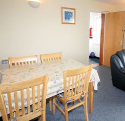 A dining area with a wooden table covered with a floral tablecloth and four wooden chairs next to a black leather sofa in a room at Polzeath Court 4 in Polzeath