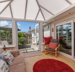 A conservatory with a sofa and cushions a wooden chair with cushions a round red rug and patio doors leading to an outdoor deck with a table umbrella at Cogenhoe in Rock