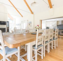 A dining room with a wooden table and white chairs next to an open kitchen with white cabinets and stainless steel stove at Ragleighs in Daymer Bay