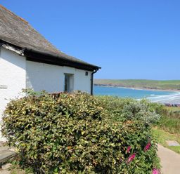 Exterior view of a white cottage with bushes in front and a beach with waves and green hills in the background at Gullsway Kittiwake in Polzeath
