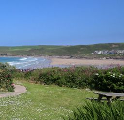 A grassy area with bushes and a wooden picnic table overlooking a sandy beach and ocean with hills and buildings in the background at Gullsway September Tide in Polzeath