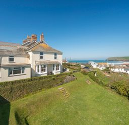 A white house with chimneys on a hill overlooking a grassy yard and coastal village at Pentire View 3 in Polzeath