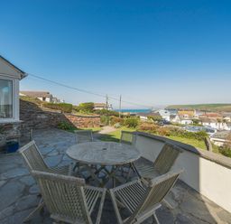 A stone patio with a round wooden table and six chairs overlooking houses and the sea at Pentire View (3) in Polzeath