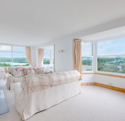 A living room with cream sofas, large windows with curtains, and views of hills and houses at Pentire View (3) in Polzeath
