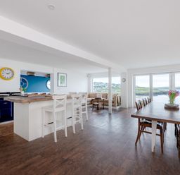 An open kitchen and dining area with wooden floors and a large table with chairs near windows overlooking a coastal landscape at Treverden Polzeath