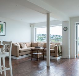 A living room with a beige corner sofa and wooden coffee table by a large window and glass door showing a coastal view at Treverden in Polzeath