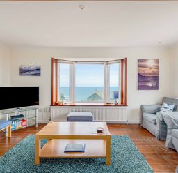 A living room with a wooden coffee table a blue rug two grey armchairs a blue chair a flat screen TV and a large window with sea view at Shilling Stones in Port Isaac