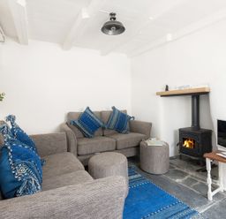 A living room with two gray sofas with blue cushions a wood stove a small wooden table with a television and a blue rug at Bre Cottage in Port Isaac