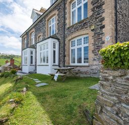 An outdoor area with a picnic table on grass next to a stone house with multiple windows and a stone wall with greenery at Folly 2 in Port Isaac