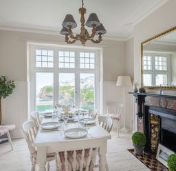 A dining room with a white table set for six by a window overlooking water and cliffs at Folly 2 in Port Isaac