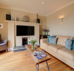 A living room with a beige sofa and blue pillows a wooden coffee table with magazines and flowers a fireplace with a mounted TV and a wicker chair at Worthy House in Daymer Bay