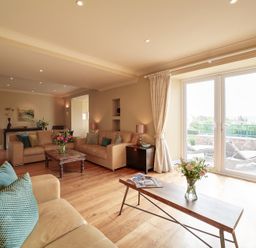 A living room with beige sofas wooden tables flowers and glass doors leading to an outdoor area at Worthy House in Daymer Bay