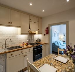 A kitchen with white cabinets wooden countertops a stove and a sink next to a dining table with a floral tablecloth and flowers at April Cottage in Port Isaac