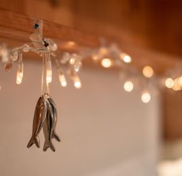 Two metallic fish ornaments hanging from a string attached to wooden beam with string lights in the background at April Cottage in Port Isaac